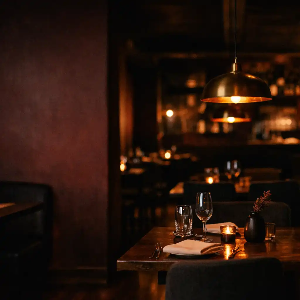 Low-lit Riverand Cinder dining room with dark timber tables, a set table and warm pendant lighting.